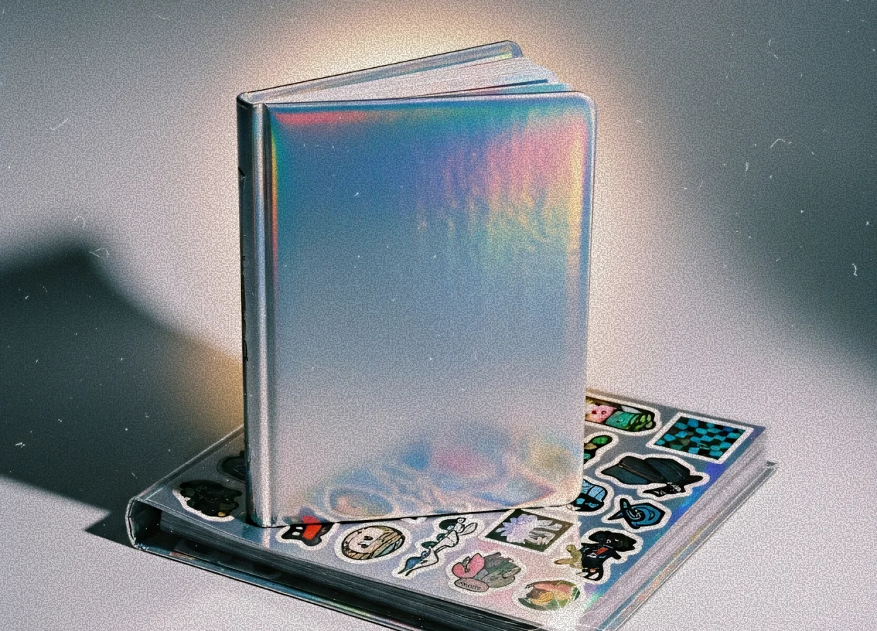 A reflective, iridescent notebook standing upright on top of a sticker-covered binder on a light background.