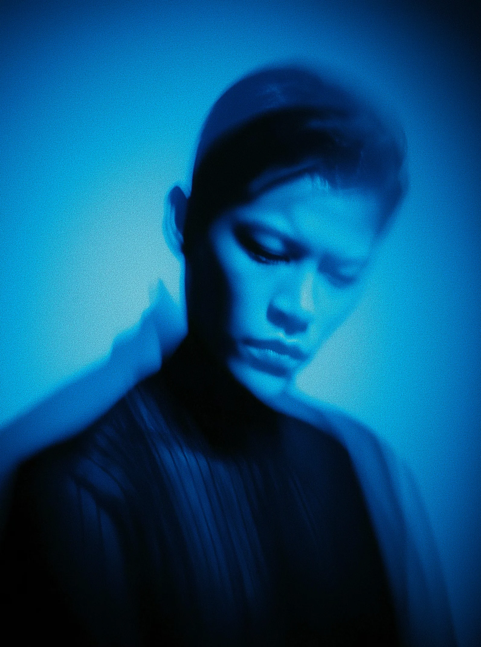 Blurred close-up portrait of a person with short hair in blue lighting, looking down with a serious expression.